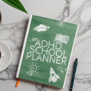 Może przedstawiać: Zielony planer szkolny ADHD z białym tekstem i ilustracjami, w tym czapką absolwenta, otwartą książką, żar&oacute;wką i rakietą. Planer ma biały grzbiet i pomarańczową wstążkę do zakładki. Pi&oacute;ro spoczywa obok planera na marmurowej powierzchni.