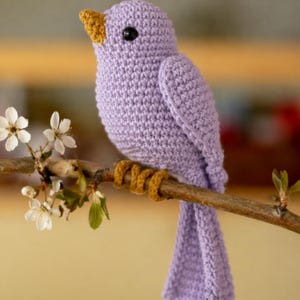May include: A crocheted lavender bird with a yellow beak and eye, perched on a brown branch with white flowers. The bird's feet are a golden-brown color. The bird is a handmade craft item.