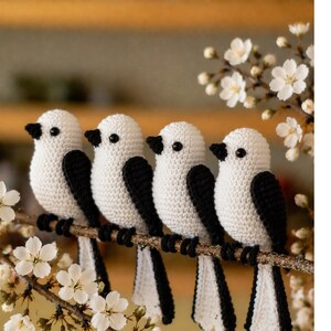 May include: Four handmade crochet birds with white bodies, black wings and tails, and black beaks are perched on a brown branch. The birds are arranged in a row, with white flowers in the foreground and background.