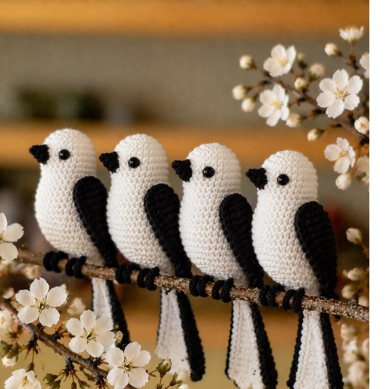 May include: Four handmade crochet birds with white bodies, black wings and tails, and black beaks are perched on a brown branch. The birds are arranged in a row, with white flowers in the foreground and background.