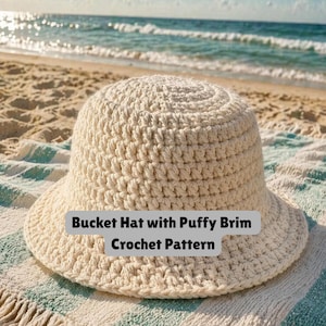 Puede incluir: Un sombrero de cubo de ganchillo de color crema con un ala abombada. El sombrero está sobre una toalla a rayas en una playa de arena con el océano al fondo. El texto "Bucket Hat with Puffy Brim Crochet Pattern" se muestra en la imagen.