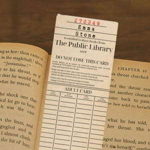 Może przedstawiać: Karta biblioteczna w stylu vintage z nazwiskiem "Emma Stone" i numerem "270349". Karta jest włożona do otwartej książki. Na karcie widnieje napis "The Public Library" i "Adult Card".