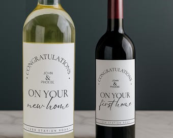 Puede incluir: Dos botellas de vino, una con vino blanco y otra con vino tinto. Ambas botellas tienen etiquetas blancas con el texto "FELICIDADES" y "ON YOUR new home" o "ON YOUR first home". Las etiquetas también incluyen los nombres "JOHN & PHOEBE" y una dirección.