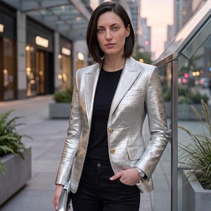 Pode incluir: Um blazer prateado metalizado com textura de pele de crocodilo, com lapelas entalhadas, dois botões dourados e bolsos com aba. O blazer é usado sobre uma t-shirt preta lisa e calças pretas, com uma pequena bolsa de mão prateada na mão.