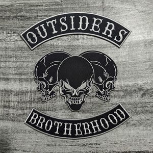 Puede incluir: Parches bordados en blanco y negro con las palabras "OUTSIDERS" y "BROTHERHOOD". Los parches presentan tres diseños de calaveras. Los parches están sobre una superficie de madera gris.