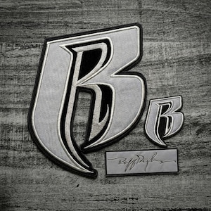 Può includere: Tre toppe ricamate con la lettera "B" in un carattere gotico stilizzato. La toppa più grande è grigio chiaro con accenti neri e bianchi. Sono inclusi anche una toppa "B" più piccola e un'etichetta rettangolare con il nome.