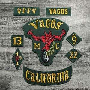 Puede incluir: Un conjunto de parches bordados en tonos verdes y dorados. Los parches presentan textos como "VAGOS" y "CALIFORNIA", junto con un gráfico de diablo rojo y los números 13, 6 y 22. Los parches están dispuestos sobre una superficie gris con vetas de madera.