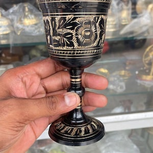 May include: Black goblet with intricate floral and geometric etched designs. The goblet has a flared base and a slender stem with gold-colored accents. The rim and base feature a decorative border. The goblet is held in a hand.