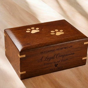 Könnte beinhalten: Eine rechteckige Urnenbox aus dunkelbraunem Holz für Haustiere mit zwei hellen Pfotenabdruck-Einlagen auf dem Deckel. Die Vorderseite ist mit folgendem Text graviert: IN LOVING MEMORY OF A Loyal Companion Forever in our Hearts 2015-2023.