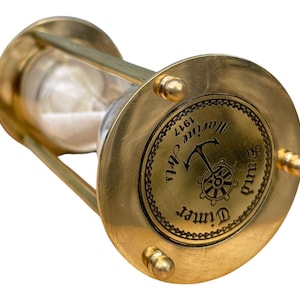 May include: A brass hourglass with a nautical design. The timer has a clear glass chamber filled with white sand. The base features an anchor and ship's wheel design with the words "Marine Arts" and "Sand Timer 1917".