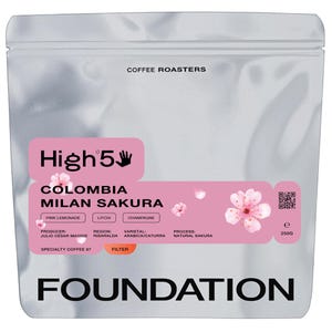 May include: A silver coffee bag with a pink label featuring the text "High 5" and "Colombia Milan Sakura." The bag includes the text "Coffee Roasters" and "Foundation." The bag contains 250g of coffee.