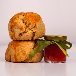 K&ouml;nnte beinhalten: Zwei goldbraune Scones, &uuml;bereinander gestapelt, mit einem kleinen Glas roter Marmelade daneben. Das Glas hat einen goldenen Deckel und ein gr&uuml;nes Band. Die Scones haben eine leicht raue Textur.