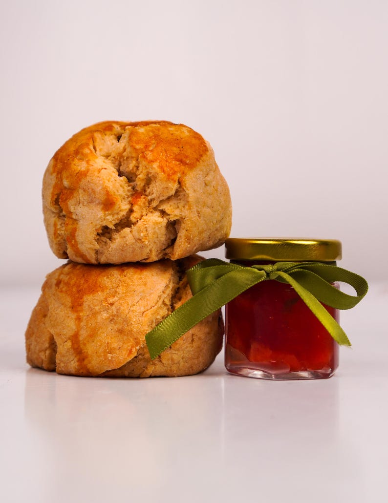 K&ouml;nnte beinhalten: Zwei goldbraune Scones, &uuml;bereinander gestapelt, mit einem kleinen Glas roter Marmelade daneben. Das Glas hat einen goldenen Deckel und ein gr&uuml;nes Band. Die Scones haben eine leicht raue Textur.