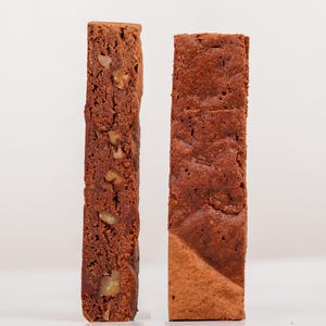 May include: Two rectangular slices of chocolate cake, one with visible nuts. The cakes are a rich brown colour and stand upright against a white background. The texture of the cake is visible, suggesting a moist interior.