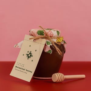 May include: A jar of botanical honey with a floral patterned lid and a tag that reads "Botanical honey". A wooden honey dipper rests beside the jar. The jar is on a red surface against a pink background.