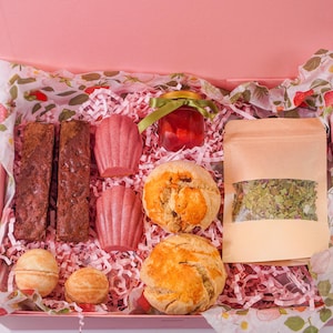 K&ouml;nnte beinhalten: Eine rosa Geschenkbox gef&uuml;llt mit Backwaren und Tee. Die Box enth&auml;lt zwei Brownies, zwei rosa Madeleines, zwei Scones, ein Glas Marmelade, einen Teebeutel und zwei Badebomben. Die Box ist mit rosa Papier und Papier mit Erdbeermuster ausgekleidet.
