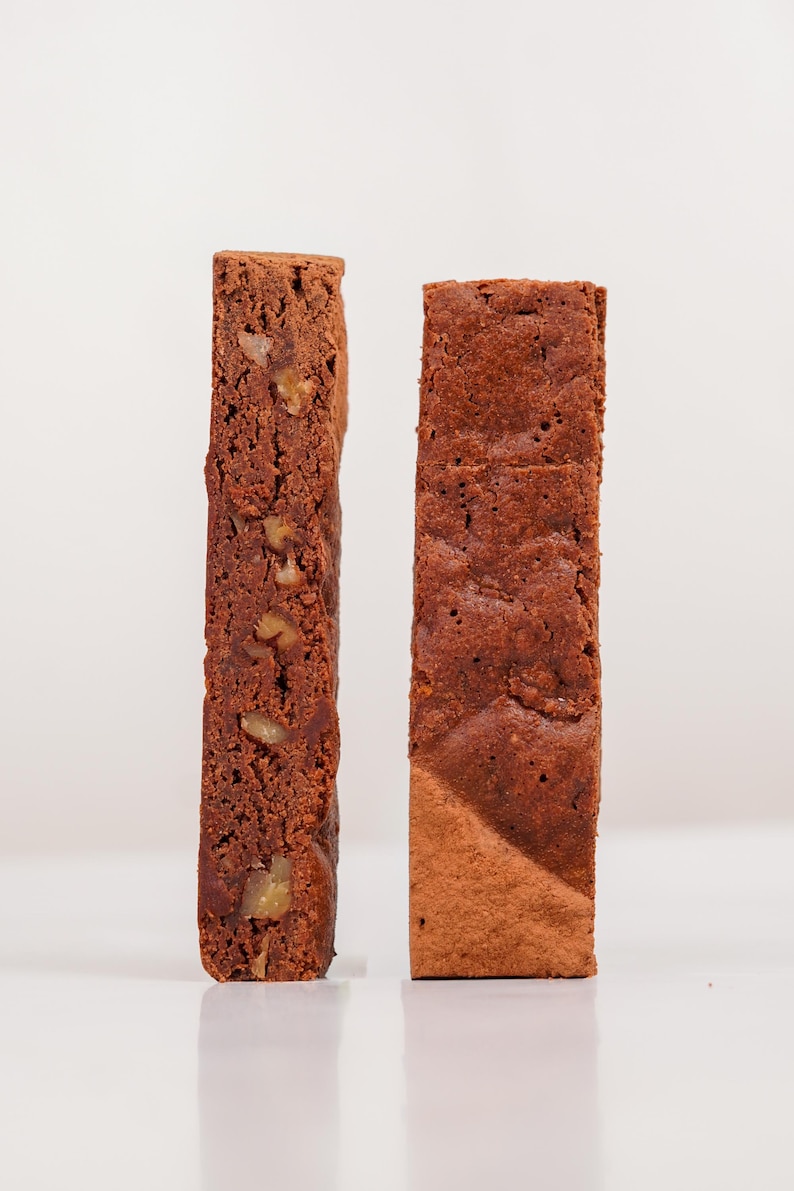 K&ouml;nnte beinhalten: Zwei rechteckige Scheiben Schokoladenkuchen, eine mit sichtbaren N&uuml;ssen. Die Kuchen haben eine satte braune Farbe und stehen aufrecht vor einem wei&szlig;en Hintergrund. Die Textur des Kuchens ist sichtbar, was auf ein feuchtes Inneres hindeutet.