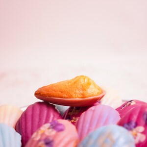 K&ouml;nnte beinhalten: Nahaufnahme einer Sammlung bunter Madeleines. Die Madeleines sind in verschiedenen Farben erh&auml;ltlich, darunter Orange, Rosa, Blau und Wei&szlig;. Einige sind mit essbaren Blumen verziert. Die Madeleines sind auf einem Teller angeordnet.