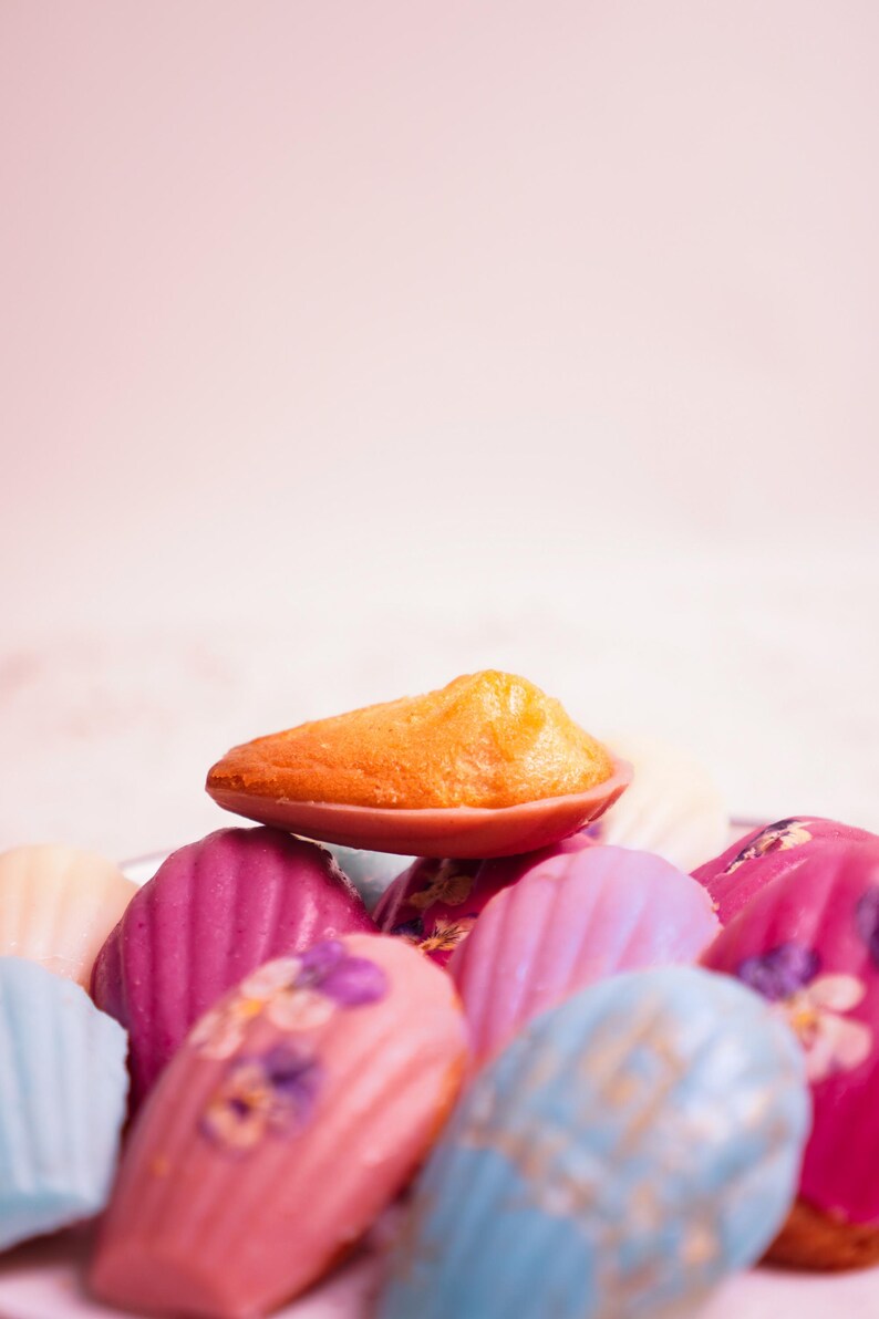 K&ouml;nnte beinhalten: Nahaufnahme einer Sammlung bunter Madeleines. Die Madeleines sind in verschiedenen Farben erh&auml;ltlich, darunter Orange, Rosa, Blau und Wei&szlig;. Einige sind mit essbaren Blumen verziert. Die Madeleines sind auf einem Teller angeordnet.