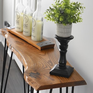 May include: A wooden console table with black hairpin legs. Three clear glass bottles with white candles sit on a wooden tray. A small potted plant in a gray and white striped pot sits on a black candlestick.