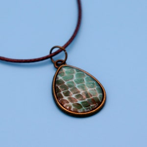 May include: A teardrop-shaped pendant necklace with a green and brown snakeskin pattern. The pendant is set in a bronze-colored metal frame and hangs from a brown leather cord.