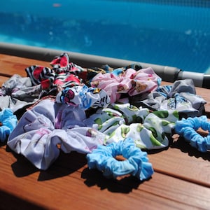 Può includere: Una collezione di elastici per capelli colorati con vari motivi e colori, tra cui blu, rosa e floreali. Gli elastici sono disposti su una superficie di legno vicino a una piscina.