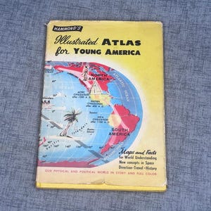 May include: A vintage "Hammond's Illustrated Atlas for Young America" book. The cover is yellow with a world map illustration in red, blue, and white. Text includes "Maps and Facts" and "Our Physical and Political World in Story and Full Color."