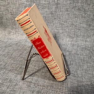 May include: A vintage book titled "Ohne Mütter geht es nicht" in German, displayed on a silver metal book stand. The book has a beige cover with red and blue accents and a red label.