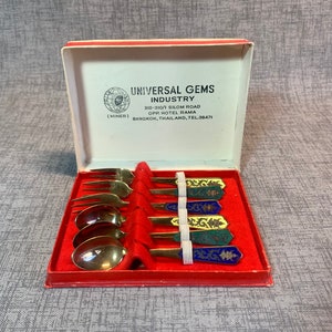 May include: A set of six gold-colored teaspoons with colorful enamel handles in a red box. The box has a label that reads "Universal Gems Industry 310-310/1 Silom Road Opp Hotel Rama (Miner) Bangkok, Thailand, Tel. 38471".
