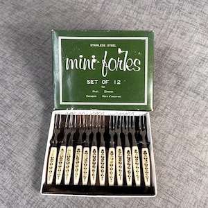 May include: A set of 12 stainless steel mini forks for fruit, cheese, canapés, and hors d'oeuvres. The forks have white handles with gold floral designs. The forks are in a green box with the words "mini forks" printed on the front.