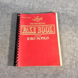 May include: Red spiral bound book with the title "Legit Professional Fake Book More Than 1010 Songs" in gold lettering. The book is by Richard Wolfe.