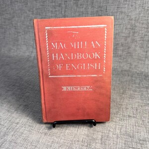 Könnte beinhalten: Ein rotes Vintage-Buch mit dem Titel "Macmillan Handbook of English" mit weißer Schrift. Das Buch steht auf einem schwarzen Metallständer. Der Name des Autors, "Kierzek", ist am unteren Rand des Buchcovers aufgedruckt.