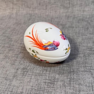 May include: A white ceramic egg-shaped trinket box with a colorful floral and bird design. The box features a bright orange and purple bird with a long tail, surrounded by pink, yellow, and green flowers.