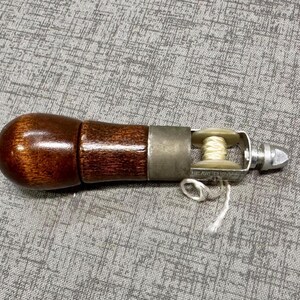 Puede incluir: Un mango de madera marr&oacute;n con un punz&oacute;n de metal y una peque&ntilde;a bobina de hilo blanco. El punz&oacute;n se utiliza para perforar agujeros en cuero y otros materiales.