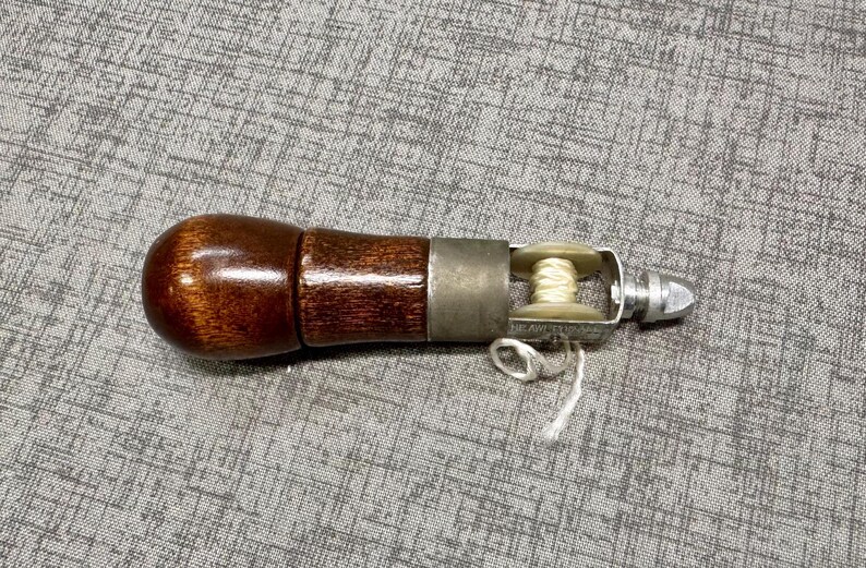 Puede incluir: Un mango de madera marr&oacute;n con un punz&oacute;n de metal y una peque&ntilde;a bobina de hilo blanco. El punz&oacute;n se utiliza para perforar agujeros en cuero y otros materiales.