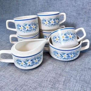 May include: A collection of ceramic cups and a creamer with a floral design. The cups and creamer are white with blue and yellow floral patterns and blue trim. The set is arranged on a blue surface.