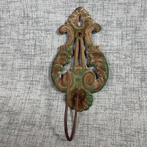 May include: A wall-mounted metal hook featuring an ornate, symmetrical scrollwork design with a weathered finish. The piece displays a combination of rusted brown and faded green paint, with a curved hook extending from the bottom section.