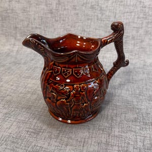 May include: A brown ceramic pitcher with a detailed relief design. The pitcher has a decorative handle and spout. The relief depicts figures and heraldic symbols. The pitcher is approximately 8 inches tall.