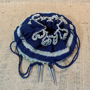 May include: A navy blue crocheted drawstring bag with silver beads and white trim. The bag has three silver metal tassels.