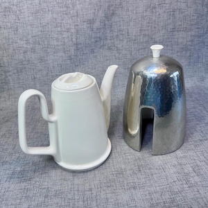 May include: A white ceramic teapot with a curved handle and spout, next to a silver metal sugar bowl with a hammered texture. The sugar bowl has a vertical opening and a small white knob on top.
