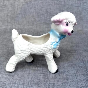 May include: A white ceramic lamb planter with a light blue scarf and pink accents on the ears and cheeks. The lamb has black eyes and a black nose. The planter has a hollowed-out back for holding small plants or decorations.