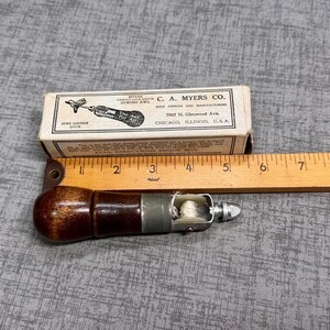 Puede incluir: Un punz&oacute;n de coser de cuero vintage con un mango de madera y una aguja de metal. El punz&oacute;n est&aacute; en una peque&ntilde;a caja de cart&oacute;n con el texto "C.A. Myers Co. 2007 N. Glenwood Ave. Chicago, Illinois, U.S.A." impreso en ella.