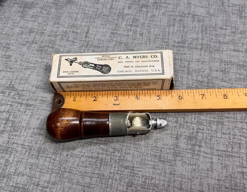 Puede incluir: Un punz&oacute;n de coser de cuero vintage con un mango de madera y una aguja de metal. El punz&oacute;n est&aacute; en una peque&ntilde;a caja de cart&oacute;n con el texto "C.A. Myers Co. 2007 N. Glenwood Ave. Chicago, Illinois, U.S.A." impreso en ella.