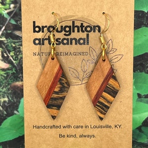 May include: A pair of diamond-shaped wooden earrings with a red, brown, and black wood grain pattern. The earrings are handcrafted with care in Louisville, KY. The earrings are attached to gold-colored hooks.