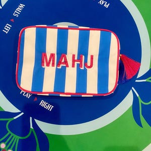 May include: A rectangular pouch with blue and white vertical stripes, red trim, and the embroidered word "MAHJ" in pink. A pink and blue tassel hangs from the side. The pouch is on a blue and green surface with text that says "PLAY RIGHT" and "WALLS LEFT".