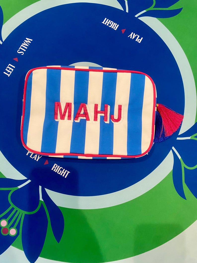 May include: A rectangular pouch with blue and white vertical stripes, red trim, and the embroidered word "MAHJ" in pink. A pink and blue tassel hangs from the side. The pouch is on a blue and green surface with text that says "PLAY RIGHT" and "WALLS LEFT".