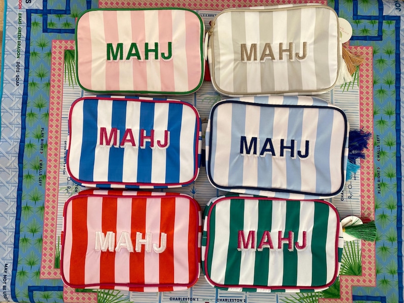 May include: Six rectangular cosmetic bags with vertical stripes in various colors: pink, gray, blue, red, and green. Each bag has the embroidered letters "MAHJ" in a contrasting color. The bags are arranged on a patterned surface.