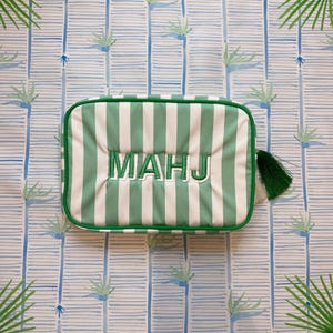 May include: A rectangular, green and white striped bag with the embroidered word "MAHJ" in white. The bag has a green tassel and is set against a patterned background with palm tree and bamboo designs. The text "OH MY MAHJONG" is visible.