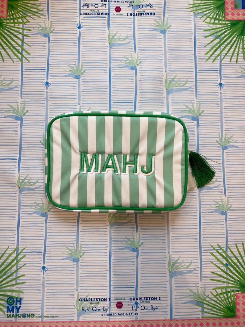 May include: A rectangular, green and white striped bag with the embroidered word "MAHJ" in white. The bag has a green tassel and is set against a patterned background with palm tree and bamboo designs. The text "OH MY MAHJONG" is visible.