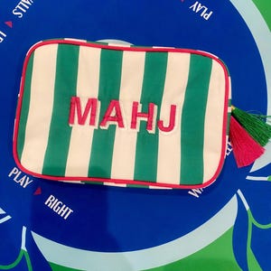 May include: A rectangular, striped pouch with green and white vertical stripes. The word "MAHJ" is embroidered in pink. The pouch has a red trim and a tassel with green and pink threads. The background is a blue and green game board.
